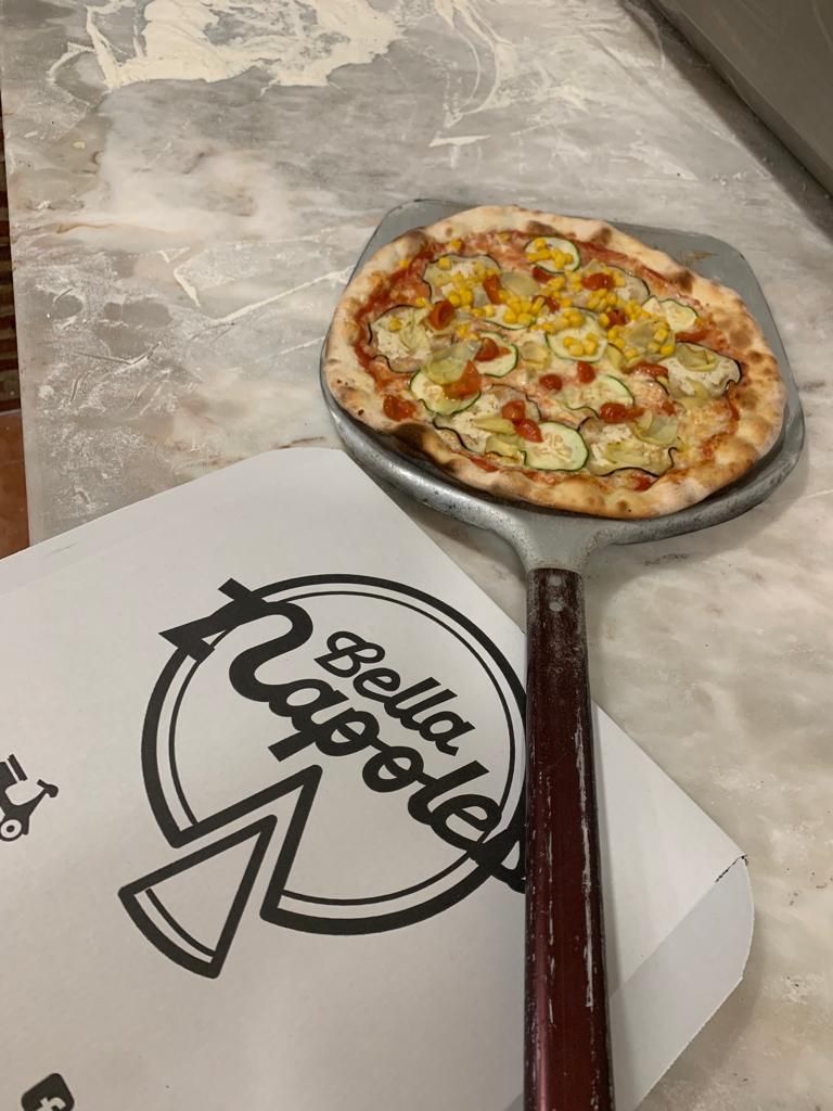Pizza de verduras con maíz recién sacada del horno en la pala, junto a una caja con el logo